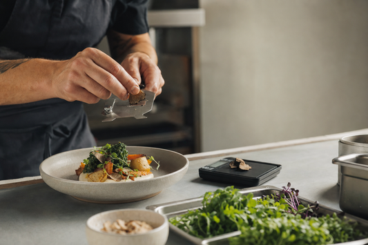 Chef applying a measured truffle garnish to a seasonal vegetable dish at the restaurant pass.